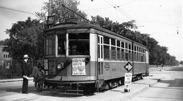 A family boarding a route #40 streetcar. A sign on the front of the car advertises midget auto racing at the State Fair grounds; the second urges people to ride and save money. The building in the background is the St. Francis Seminary.
