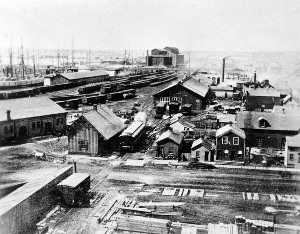 Elevated view of the first Milwaukee railroad depot, which was used by the Milwaukee & Waukesha and the Milwaukee & Mississippi railroads.  The Milwaukee & St. Paul Railway acquired the Milwaukee & Mississippi Railroad in 1868 and later diverted the passenger trains to the Union Depot on Reed Street. The presence of passenger cars to the north of the depot suggest that this picture was taken shortly prior to that move.