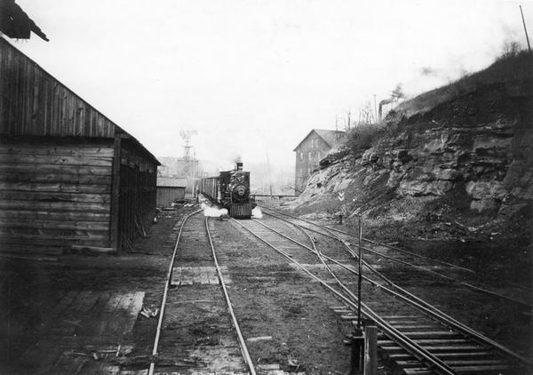 View looking down railroad tracks towards the Chicago, Milwaukee and St. Paul Railroad locomotive #240, a type 4-4-0. On the right is a steep, rocky bank, and on the left a wooden industrial building.