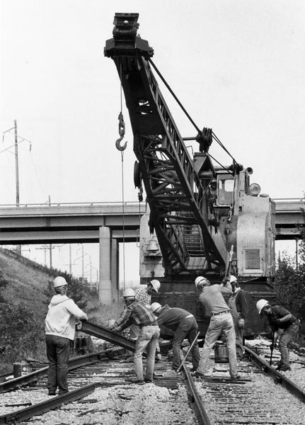 View down tracks of a repair crew working on Milwaukee Road tracks, near Rothschild, using a crane to help lift a rail. An unidentified highway bridge over the track is seen in the background.