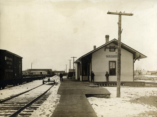 View down platform towards the Wisconsin and Michigan Railroad. A box car of the Minneapolis, Saint Paul & Sault Ste. Marie Railroad is sitting on a rail siding to the left. Snow is on the ground, and three men are standing on the platform.