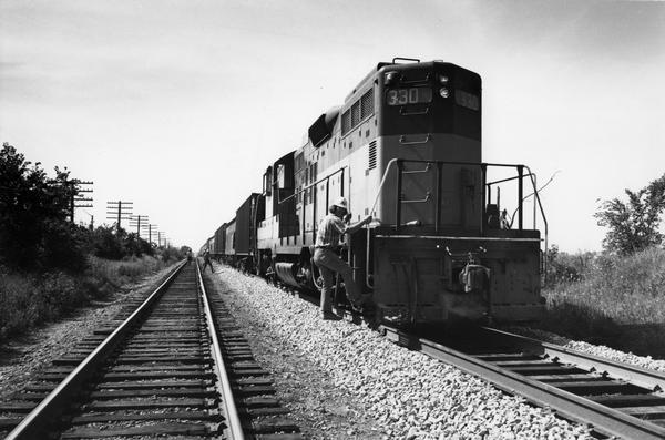 View down double set of railroad tracks towards a Milwaukee Road train, with the engineer about to board. Other men are standing in the background.