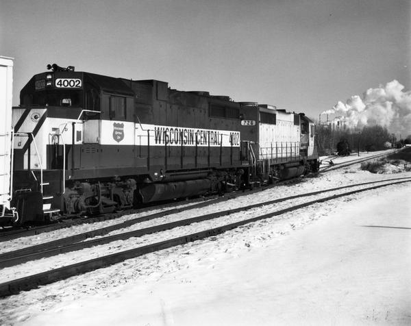 View across snow-covered railroad tracks towards Wisconsin Central's locomotive #4002. Industrial buildings are sending steam or smoke through smokestacks in the background.