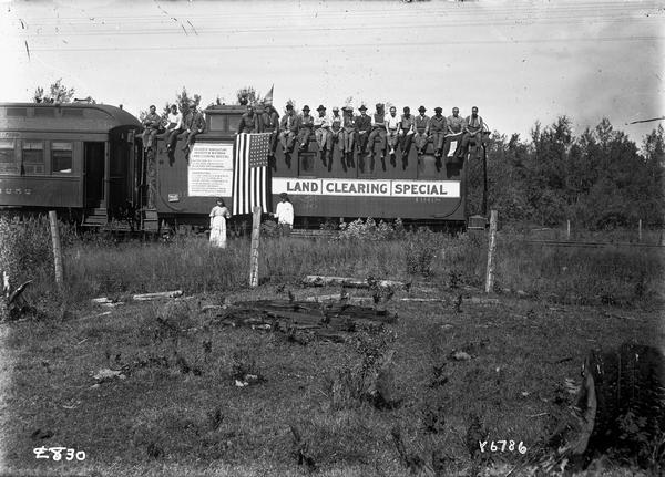View across field towards the Land Clearing Special Train, a program sponsored by the University of Wisconsin's Department of Agriculture and the U.S. Department of Agriculture. A number of men wearing work clothes are sitting on the top of a box car of the Chicago & Northwestern Railroad, which was among the corporate sponsors of the program. A man and a woman stand on the ground near an American Flag draped over the side of the box car.