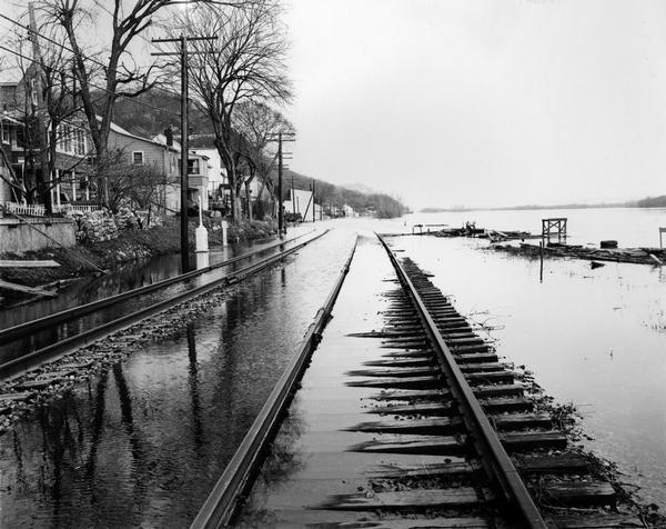 View down flooded railroad tracks of the Chicago, Burlington & Quincy Railroad by the Mississippi River. There are houses on the left, and in the background is a bluff.