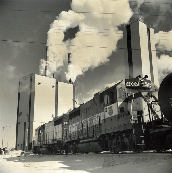 Two Wisconsin Central Railroad locomotives near the Consolidated Papers pulpmill at Wisconsin Rapids. Snow is on the ground, and heavy smoke is coming out of the smokestacks.