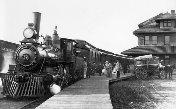 View down platform towards a locomotive pulling a Green Bay & Western Railroad train at the Green Bay depot, with a Snavely omnibus parked at the platform ready to pick up passengers for the Beaumont House.