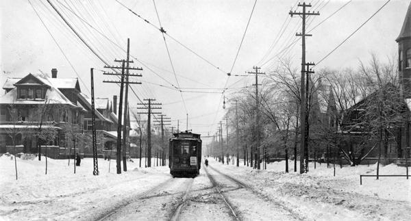 View down snow-covered streetcar tracks towards a streetcar traveling up an unidentified residential street in Milwaukee.