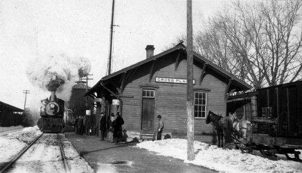 View towards railroad station with a few passengers on the platform awaiting the arrival of the train. The locomotive is billowing smoke. Two horses stand with a wagon on runners parked near the station. Snow is on the ground. A sign on the station reads, "Cross Plains."