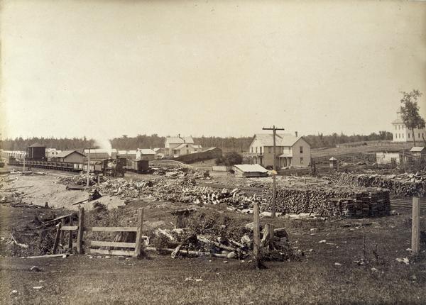 Railroad depot surrounded by stacked lumber for wood-burning locomotives. A few homes can be seen in the background.