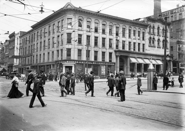 A police officer stands in the middle of the street near street railroad tracks in the pavement as pedestrians cross. In the background on the street corner is the downtown ticket office of the Chicago and Milwaukee Railroad.