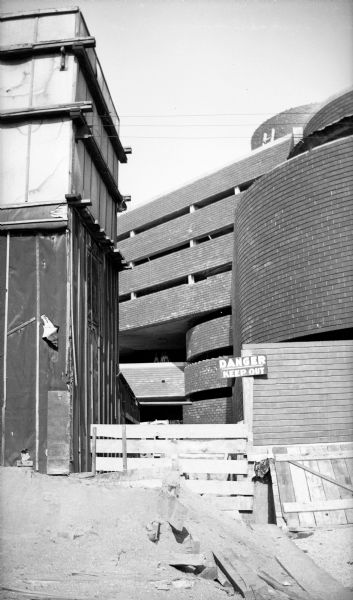 Exterior of the Johnson Wax Building during construction. The curved wall near the entrance is on the right. A construction shack is at the left. A sign posted on the construction fence states "Danger, Keep Out." The building was designed by Frank Lloyd Wright.