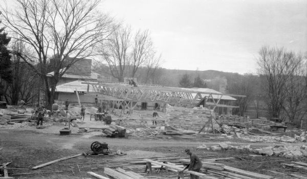 Constuction workers working on the construction of the drafting studio at the Taliesin Fellowship Complex. The roof trusses and stone walls are in place. Construction tools and supplies are scattered throughout the construction site. The studio was designed by Frank Lloyd Wright and used by the Taliesin Fellowship. Taliesin is located in the vicinity of Spring Green.