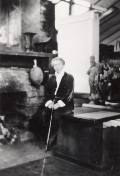 Portrait of Frank Lloyd Wright seated on a drafting table possibly in his office at Taliesin.  Wright is holding a cane and is in front of a large model of a building. Taliesin is located in the vicinity of Spring Green, Wisconsin.