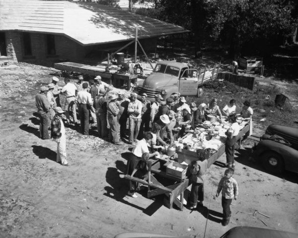 People gather around a table of food for the First Unitarian Society's "Work Day". The building was designed by Frank Lloyd Wright and some of the construction was done by members of the Meeting House congregation in order to save money. Construction workers also donated some of their time to the project.