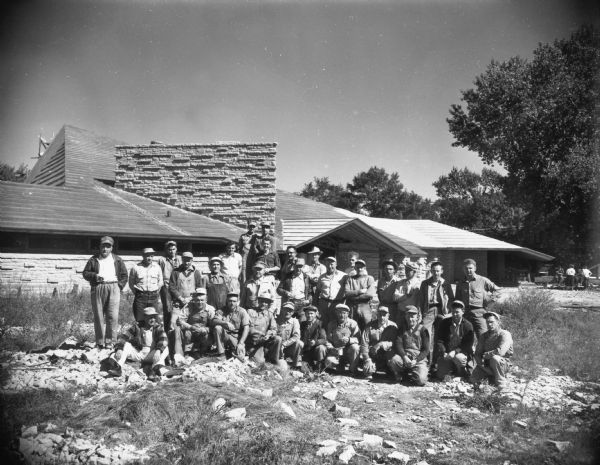 A group of skilled workmen in front of the First Unitarian Society meeting house on one of the work days they donated to the project. The building was designed by Frank Lloyd Wright. Left to right, seated: the workmen are Eathen Henstock, Milton Thorpe, Ervin Schmudlach, Chalres Manteufel, Urban Richgels, Harold Vetter, Dan Werber, Frank Tetzlaff, Howard Busse, Josephn Boykin, and Elliott Schamens. In the second row are: Guy Henry, Donald Van Schoyek, Harry Riegels, Albert Loftus, Arthur Winkler, Herbert Hulsizer, Herbert Amundson, Ralph Arneson, Erling Landsverk, Harold Johnson, Nathan Williams, and Marshall Erdman, the general contractor. In the third row are: Byron Tetzlaff, Paul Okey, Robert Shaw, Richard Gorski, Arthur Gulrud, and Lawrence Walter, and the two men in the back are William Kruse and John Johnson.
