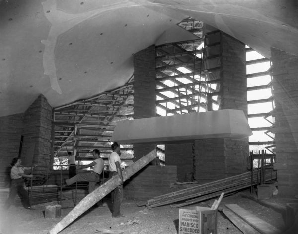 The First Unitarian Society Meeting House sanctuary (or auditorium) during the final stages of construction. Here, members of the congregation who helped during the construction in order to save money, remove planks and scaffolds. Workers are assembling or disassembling scaffolding. The building was designed by Frank Lloyd Wright and it is likely that his apprentices, who also aided in the construction, may be pictured here.