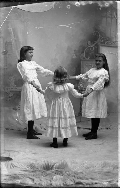 Studio portrait in front of a painted backdrop of three girls holding hands in a ring (dancing).