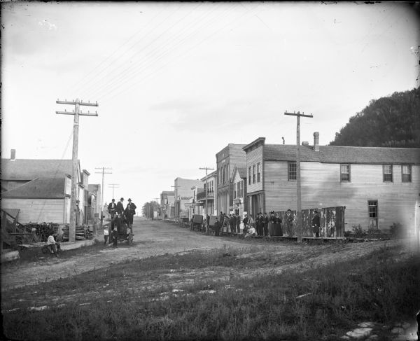 Small town business district with a group of men and women posed on the board sidewalk on the right. Another group of men and boys are on and around a tractor on the left in the dirt road. There are storefronts with signs for a Saloon, Bakery, and Post Office.