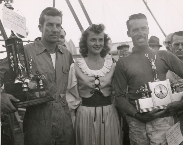 Alice in Dairyland standing with midget auto race winner Sam Hanks, a noted Indianapolis 500 driver, and Neal Carter, the fastest qualifier in the 100-mile race. Alice is wearing the special dress that was designed for her in 1948.