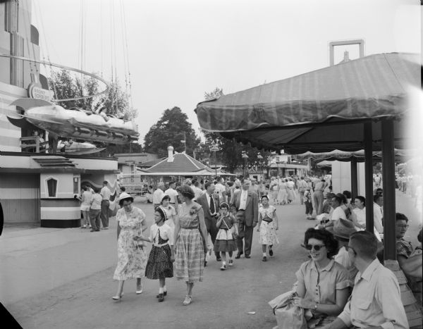 Crowds wandering among the amusement rides at the Wisconsin State Fair. In the background is the ride known as the "Rocket Bomber."