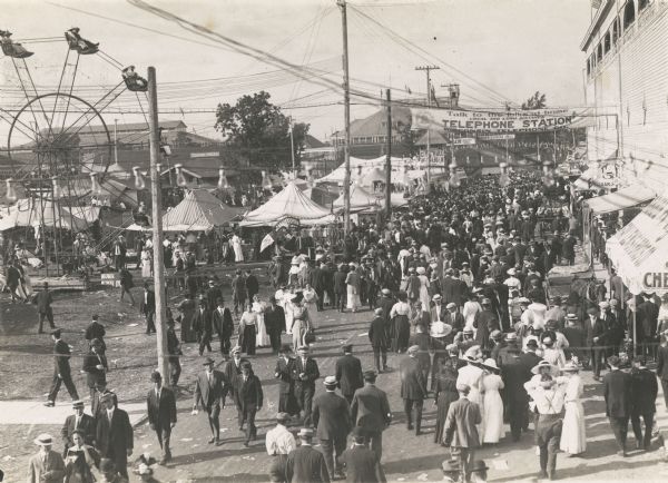 Elevated view of crowds strolling through the Wisconsin State Fairgrounds. In the background is a Ferris wheel, and a banner encouraging fair goers to "talk to the folks at home, local and long distance service, Wisconsin Telephone Co."