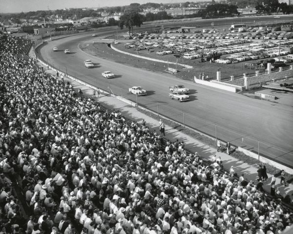 Elevated view of an automobile race at the state fairgrounds, with the crowd in the foreground. Cars are parked in the infield.