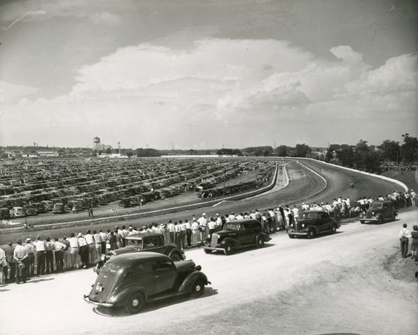 Racetrack at the Wisconsin State Fair, with a crowd lined up along the fence watching the automobile race. The infield is full of parked cars.
