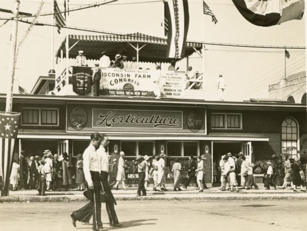 Crowds walking past the Horticulture Building at the Wiconsin State Fair.  In a pavilion above the building the Aluminum Goose Manufacturing Co. Band of Manitowoc is playing, while a banner advertises the Wisconsin Farm Congress, October 13-14, 1927, called by Governor Fred Zimmerman to address the need for farm relief.