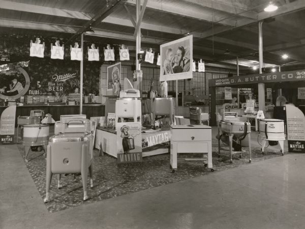Display of Maytag wringer-type washing machines in the Industry Building at the Wisconsin State Fair. In the background is a display for Miller Beer, 5 cents a glass.