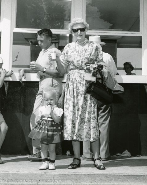 At the Department of Agriculture's Milk House at the Wisconsin State Fair, an elderly woman, Mrs. Shimmon of Milwaukee, and a young man drink milk from paper cups, while an infant, Judith Brendalen of West Allis, standing nearby drinks milk from a bottle. The Milk House was an innovative feature created in 1939 to promote milk consumption.