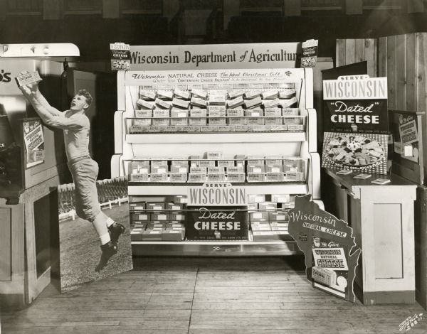 The Department of Agriculture's Cheese promotion booth at the Wisconsin Centennial Exposition. The booth urges consumers to "Serve Wisconsin Dated Cheese." The display also shows a poster of a football player catching a small sign advertising the Centennial Fair.