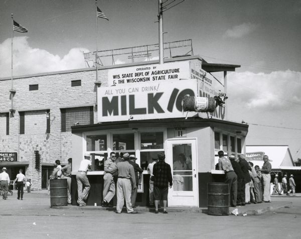 The Department of Agriculture's "All You Can Drink for Ten Cents" milk booth at the Wisconsin State Fair. On the roof is a cow crafted from a bale of hay. Miller's beer stand and the Grandstand can be seen in the background.