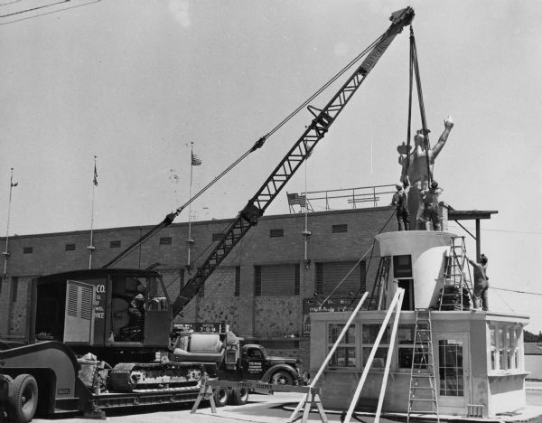 Fair employees use a crane to complete the positioning of the "Forward" statue at the Wisconsin Centennial Exposition. The grandstand can be seen in the background.