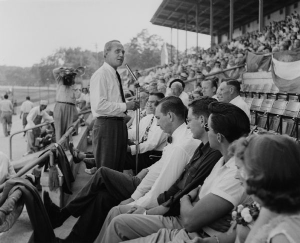 Governor Walter J. Kohler, Jr., speaks to a crowd in the grandstand at the Wisconsin State Fair. In the background, a photographer is taking his picture.