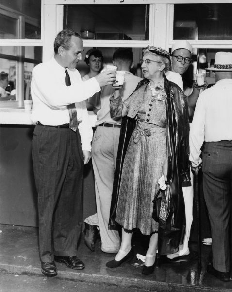Governor Walter J. Kohler, Jr., and an unidentified elderly woman, toast each other with paper cups of milk. They are standing near the Department of Agriculture's Milk House, an innovative idea introduced in 1930 to promote milk consumption.