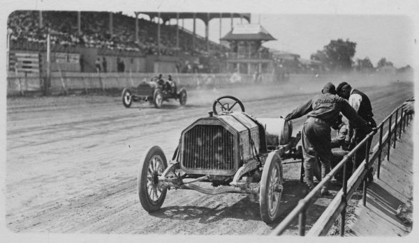 A race car driver wearing a "Buick shirt" watches as his crew works on his vehicle. In the background, another car is passing the grandstand.