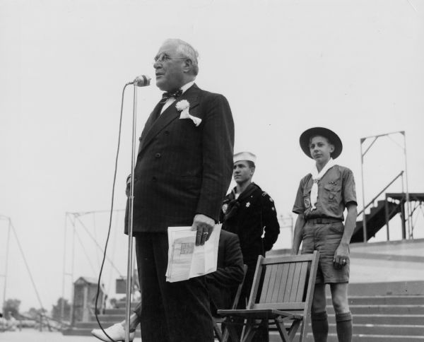 Governor Julius Heil speaks to an audience at the Wisconsin State Fair. Standing behind him are a boy scout and a U.S. Navy sailor.