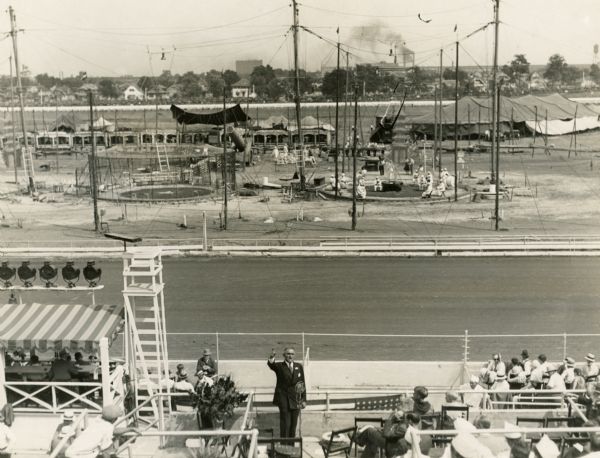 Elevated view of Governor Walter J. Kohler, Sr., speaking to an audience in the grandstand at the Wisconsin State Fairgrounds. Behind him in the infield, a circus is preparing to perform.
