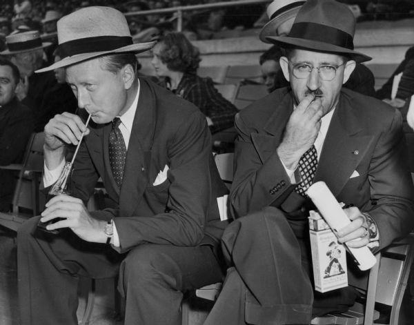 Mayor Carl Zeidler of Milwaukee and Mayor Leo J. Promen of Fond du Lac at the Wisconsin State Fair. Zeidler is sipping Coca-Cola from a bottle and Promen is eating Cracker Jacks.