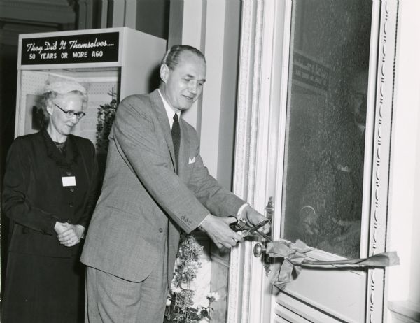 Governor Walter J. Kohler, Jr., cuts a ribbon to open the "They Did It Themselves: 50 Years Ago" exhibit at the State Historical Society. Mrs. Vincent W. Koch, SHSW Auxiliary president looks on. The exhibit opening was part of the re-dedication of the Society building after the general collection moved to the new Memorial Library and the Society building had been remodeled.