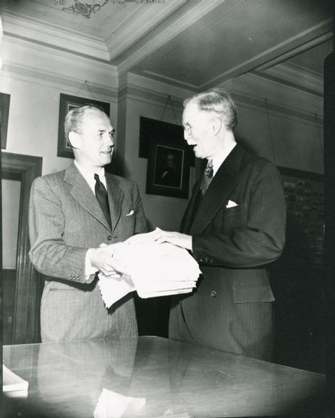 Governor Walter J. Kohler, Jr., presents a stack of papers to Secretary of State Fred R. Zimmerman. Although unidentified, the papers are likely nomination petitions. Some of the office decor can be seen, including a small portion of the original ceiling decoration.