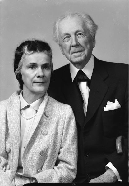 Unretouched waist-up portrait of Mr. and Mrs. Frank Lloyd Wright taken at the same time that each sat individually with Reierson for new passport photographs. Wright is holding a cane under his left arm. The photographer's original jacket says "Retouch couple picture, Mrs. Wright mostly."