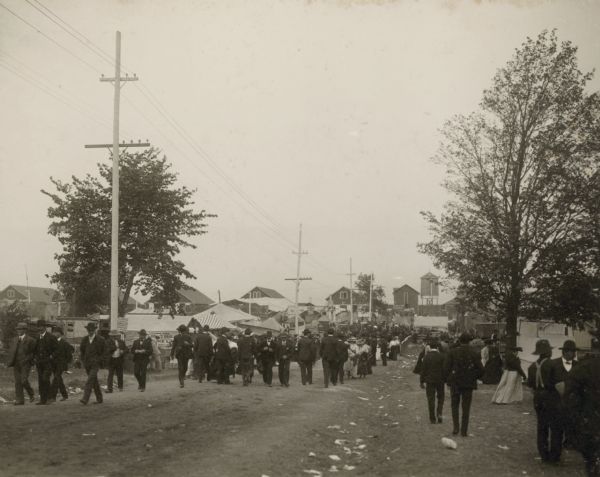 Fairgoers stroll along a dirt path within the Wisconsin State Fairgrounds. The dairy barns form a row at the back of the image.