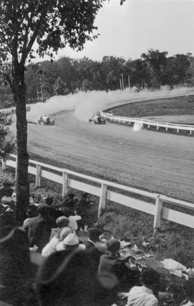 Two open cars, a Deusenberg and a Hudson, race around a curve at the Wisconsin State Fairgrounds track, spewing dust behind them. The Milwaukee Mile track at the state fair, which was not paved until 1954, is the oldest operating automobile racetrack in the nation.