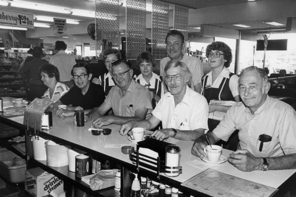 Carlene, Corey, and Laura, pose with a group of customers at the lunch counter in a Rennebohm's Drug Store restaurant. Part of the restaurant and store is visible in the background.