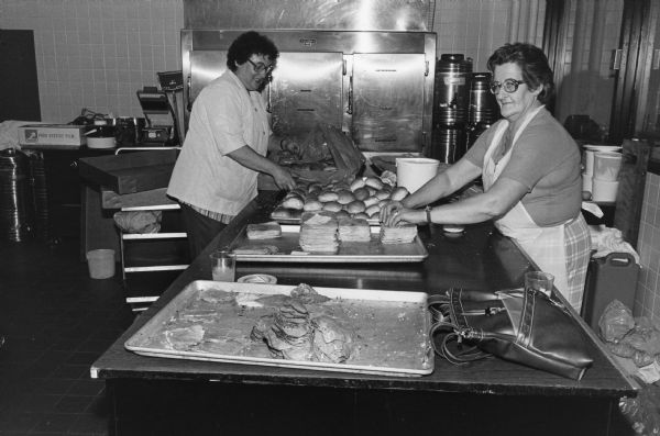 Two Rennebohm Drug Store, Inc., employees make sandwiches. A large commercial refrigerator is visible in the back of the kitchen area where they are working.