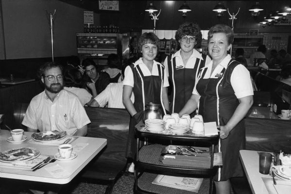 Waitresses Carlene, Laura, and Corey, pose for a photograph at an unidentified Rennebohm Drug Store restaurant. To the left, Steve Kimbrough, seated with his breakfast, looks on. Other diners can be seen in the background. Kimbrough and photographer Chuck Patch were in the midst of a project to eat breakfast at and photograph all of the existing Rennebohms.