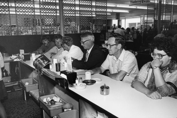 Customers sit at the lunch counter of an unidentified Rennebohm's Drug Store restaurant. The camera angle permits a view of the back of the counter, and the drug store area can be seen in the background.
