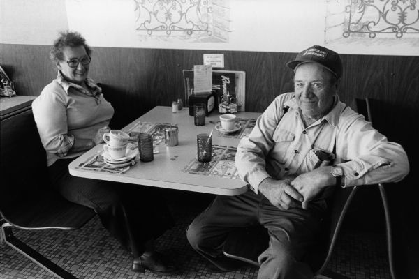An unidentified couple at an unidentified Rennebohm Drug Store restaurant. The couple had finished their meal and left their tip on the table when they were photographed by Chuck Patch. Patch was in the process of eating at and documenting all of the existing Rennebohms. The man is wearing a hat advertising Menard's.